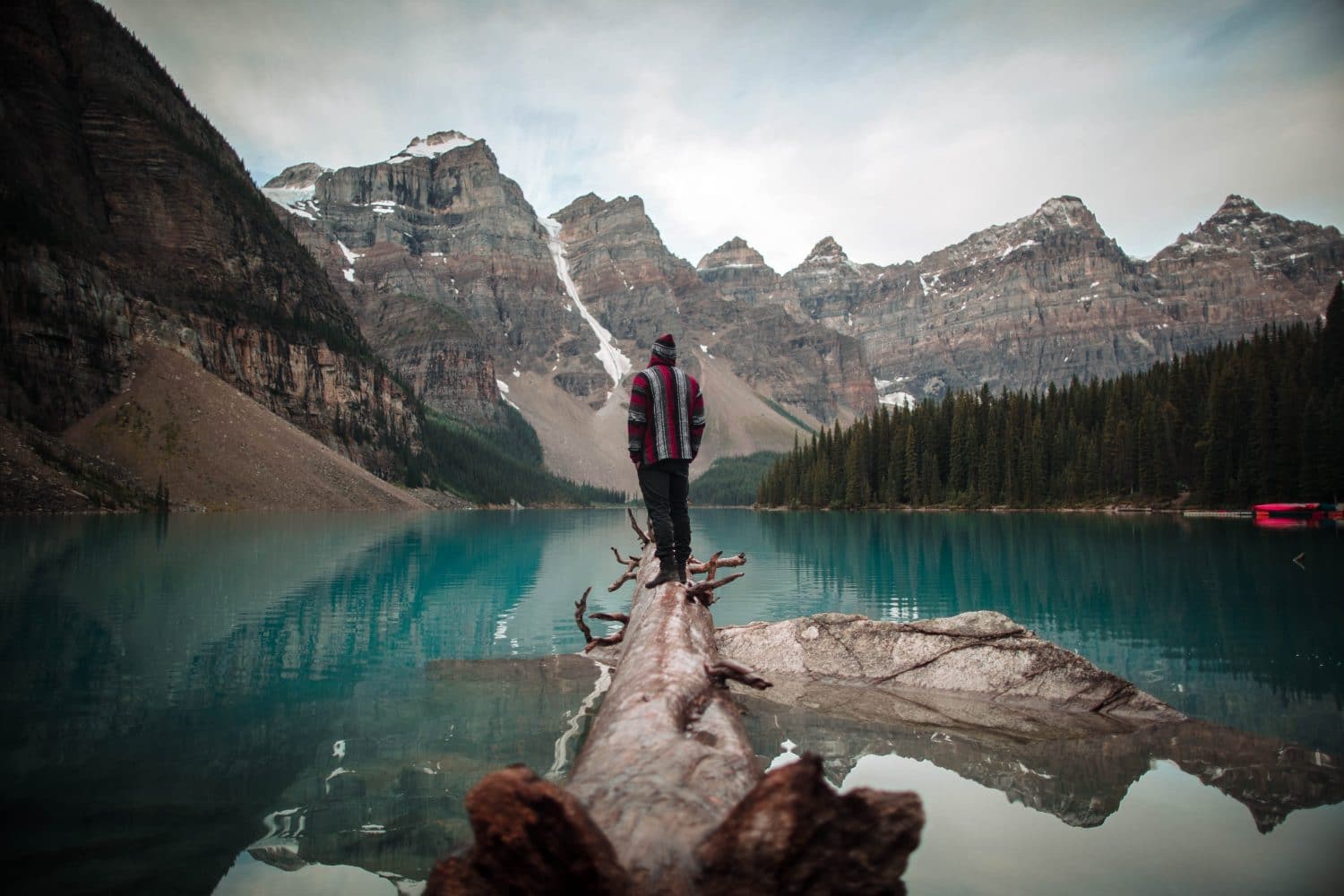 Man wearing red and white jacket standing on brown logs looking at lake and icy mountain scenery